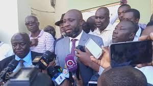 "George Afriyie addressing the media in a grey suit, surrounded by reporters and microphones."