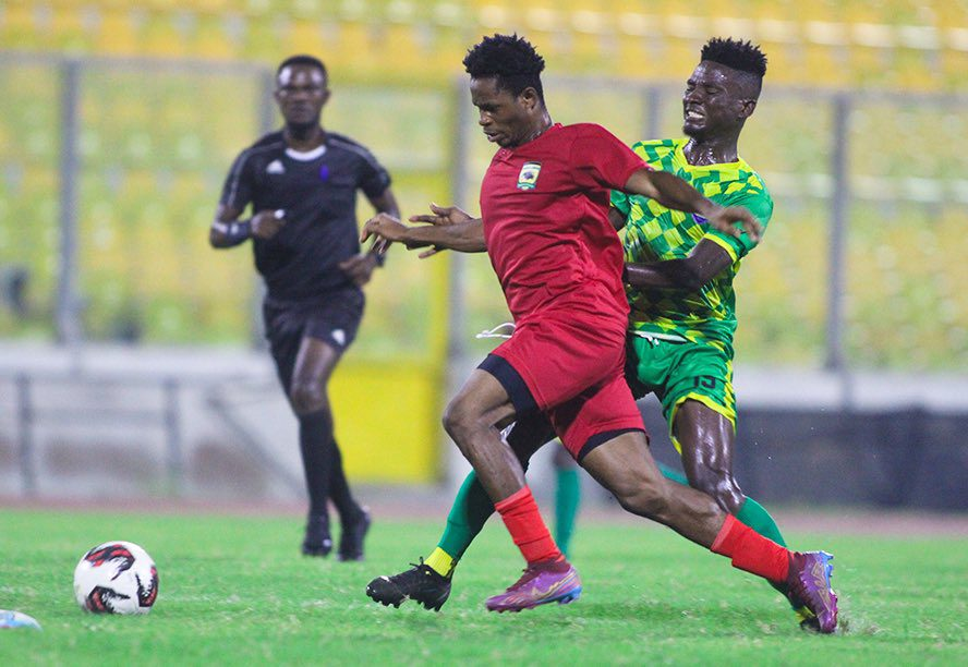 Two soccer players competing for the ball, one in red and the other in green, with a referee in the background.