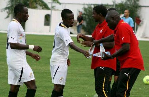 Football coach in a red shirt giving instructions to players in white kits on a grassy field during training.
