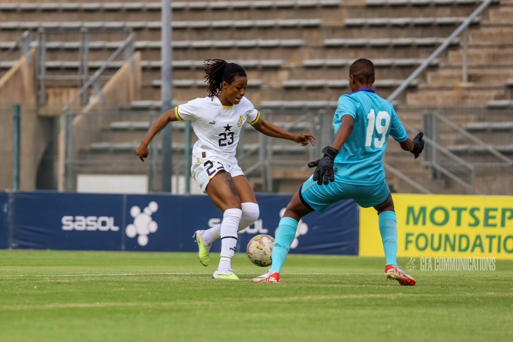 wo female soccer players in action during a match. One player, wearing a white jersey, is in possession of the ball and facing a defender in a teal jersey