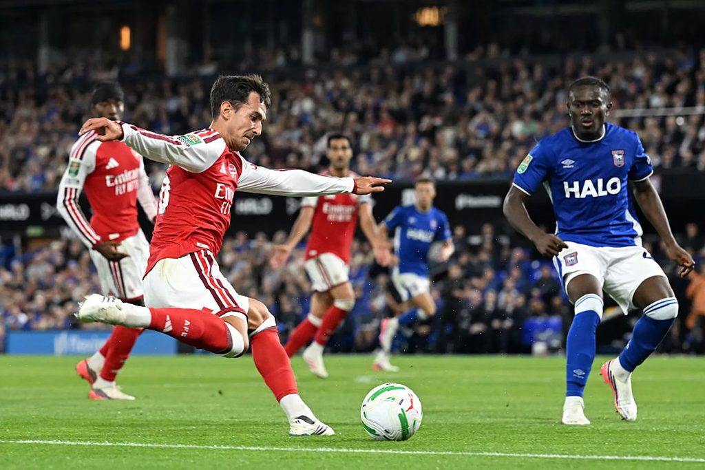Mapambano ya Mpira Uwanjani Kati ya Ipswich Town na Arsenal Mchezaji wa Arsenal akipiga mpira kwa nguvu huku akizuiliwa na beki wa Ipswich Town katika uwanja wa Portman Road