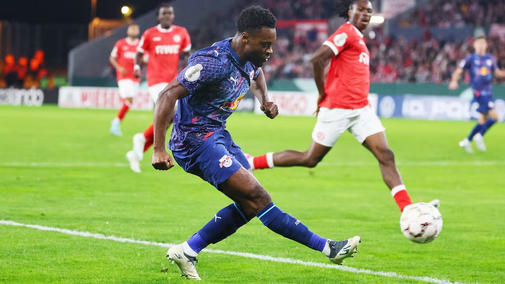 mapambano-ya-mpira-essen-vs-rb-leipzig. Mchezaji wa RB Leipzig akipiga mpira kwa kasi uwanjani huku akisindikizwa na mabeki wa Essen waliovaa jezi nyekundu.