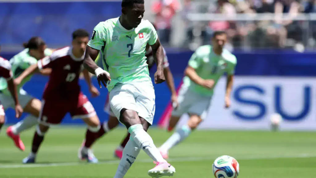 A Swiss football player in a light green kit controls the ball during a match, with opposing players in red and teammates running in the background on a sunny stadium pitch.