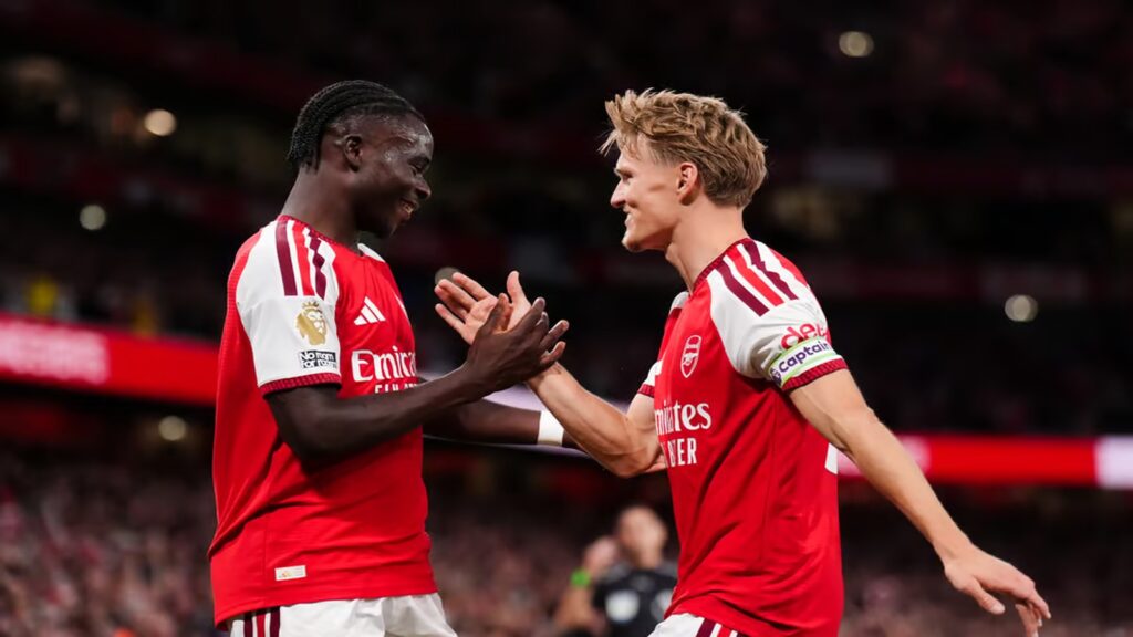 Arsenal players Bukayo Saka and Martin Ødegaard in red-and-white kits share a moment on the pitch during a football match.