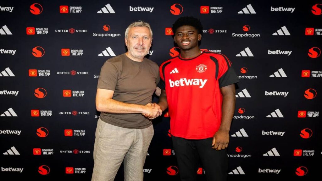 Carlos Baleba wearing a Manchester United training shirt shakes hands with a club official during his official unveiling.