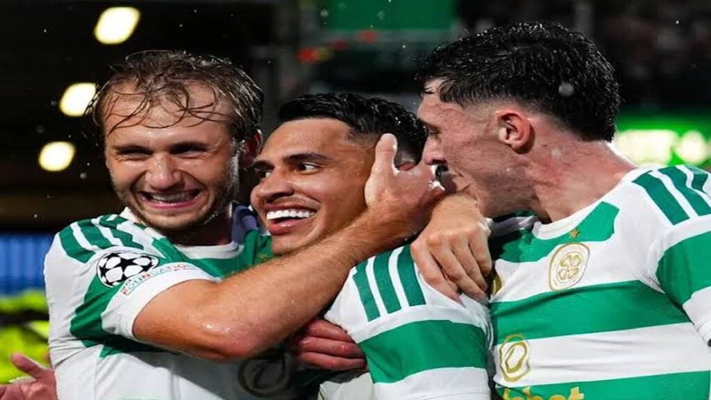 Celtic players in green-and-white kits embrace and celebrate on the pitch after scoring a goal during a football match.