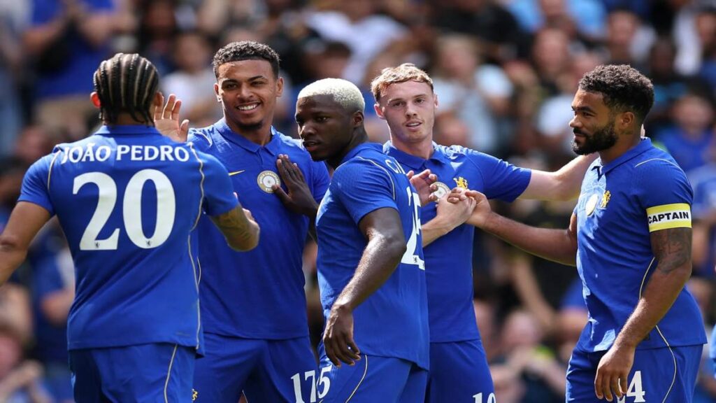 Chelsea players in blue kits gather and celebrate on the pitch after scoring, with teammates embracing during a Premier League match.