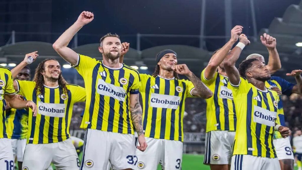 Fenerbahçe players in yellow-and-blue kits raise their arms and celebrate on the pitch after a football match.