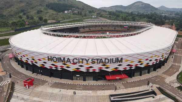 Hoima City Stadium viewed from above, showing the oval-shaped modern football venue, covered stands and surrounding landscape.