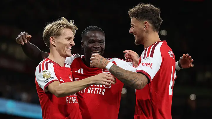 Arsenal players Martin Ødegaard, Bukayo Saka and Ben White smiling and celebrating on the pitch.