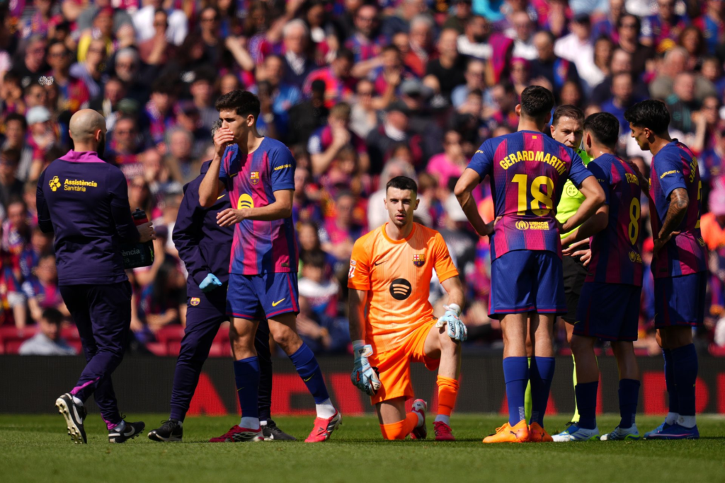 Barcelona goalkeeper Joan García kneels as teammates and match officials gather on the pitch at Camp Nou.