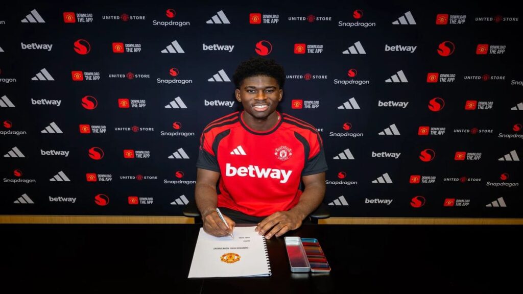 Carlos Baleba sits at a table signing his Manchester United contract while wearing a club training shirt during his official unveiling.