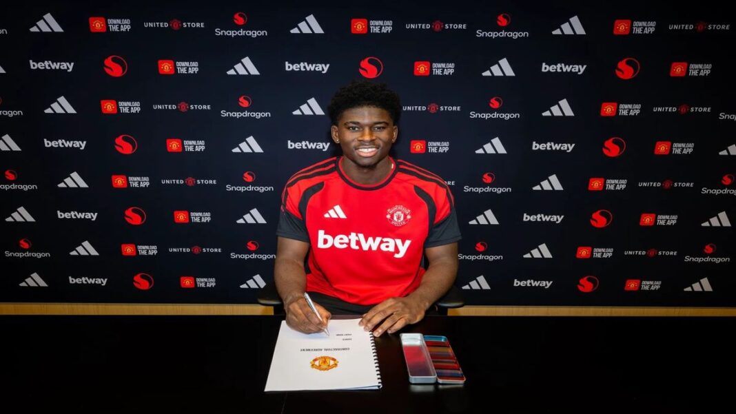 Carlos Baleba sits at a table signing his Manchester United contract while wearing a club training shirt during his official unveiling.