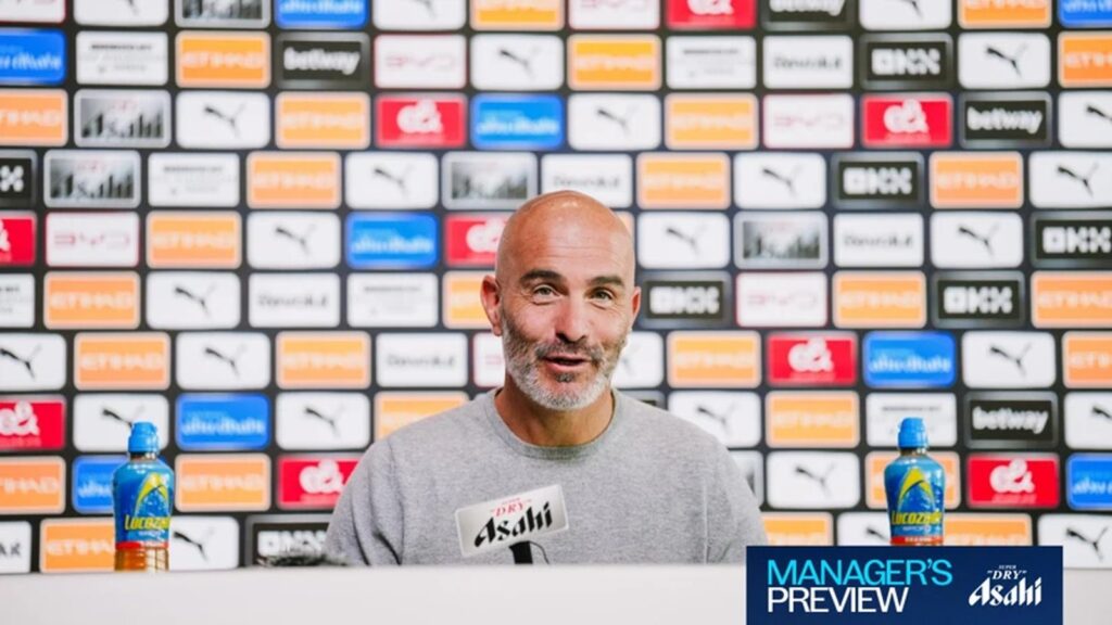 Enzo Maresca speaking at a press conference with sponsor logos displayed on the backdrop.