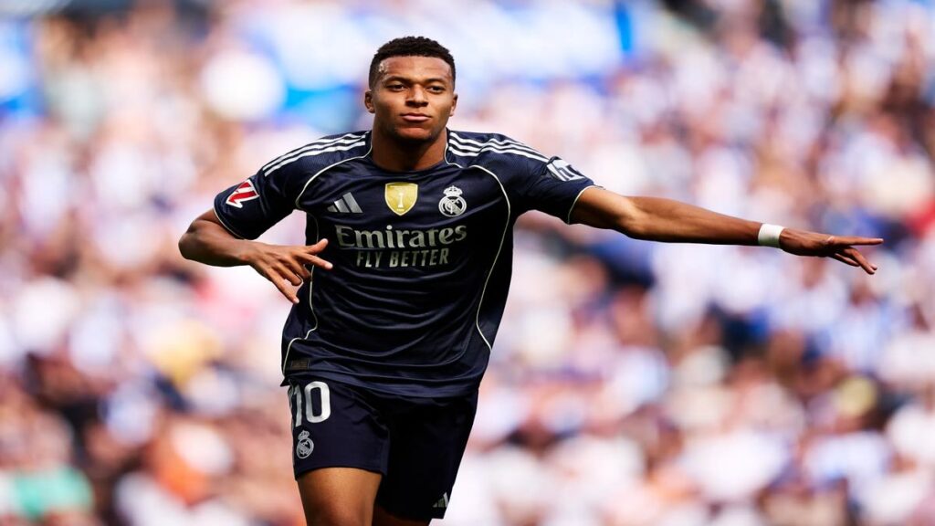 Real Madrid forward Kylian Mbappé in a dark kit celebrates with arms outstretched after scoring during a football match.