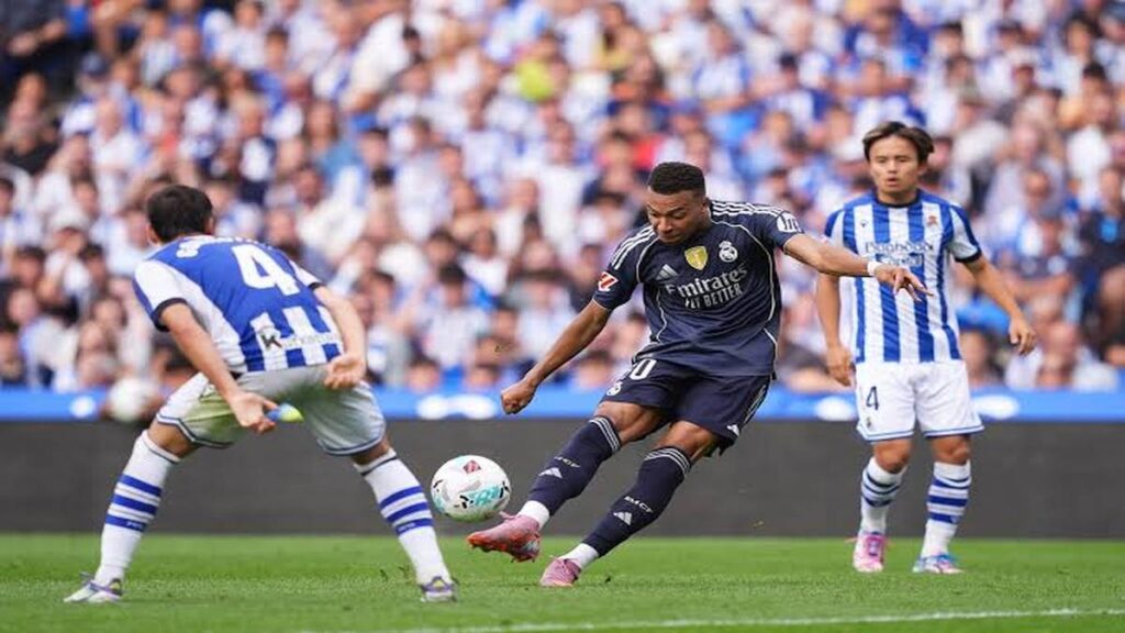 Real Madrid forward Kylian Mbappé in a dark kit takes a shot while competing against Real Sociedad defenders during a football match.