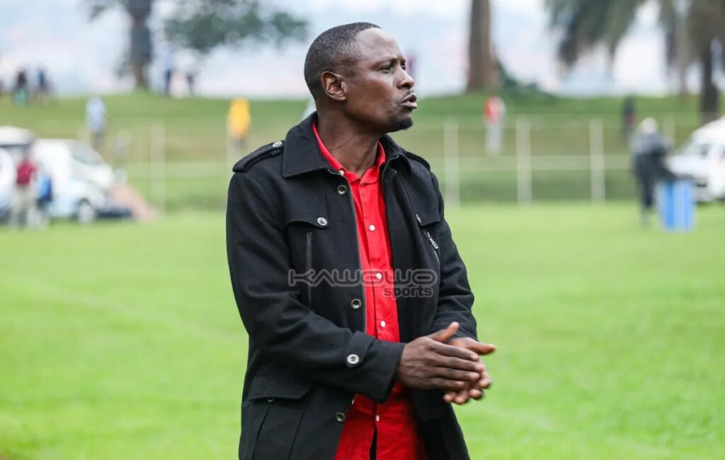 Alex Isabirye in a black jacket over a red shirt stands on a grassy sports field, hands clasped, speaking while looking off to the side; parked cars and spectators are visible in the blurred background.