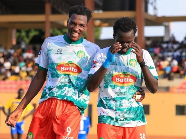 Two BUL FC footballers in green-and-white striped jerseys sponsored by Fortune Butto celebrate on the pitch — one smiling with a raised fist, the other wiping his face — with a stadium crowd visible in the blurred background.