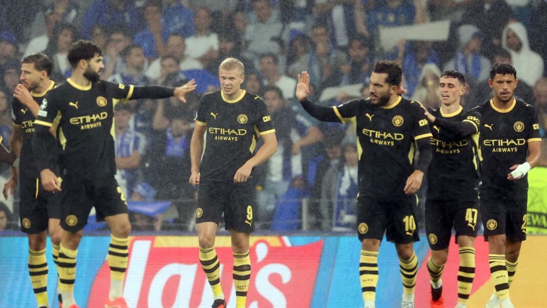 Manchester City players in black-and-gold away kits walk together on a rain-soaked pitch, one player raising a hand for a high-five with a teammate, with a large crowd filling the stands behind them.