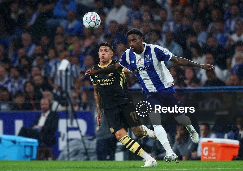 Manchester City's Enzo Fernández, in black-and-gold kit, and FC Porto's Pablo Rosário, in blue-and-white kit, jump together challenging for a header, with a packed stadium crowd blurred behind them and a Reuters watermark visible.