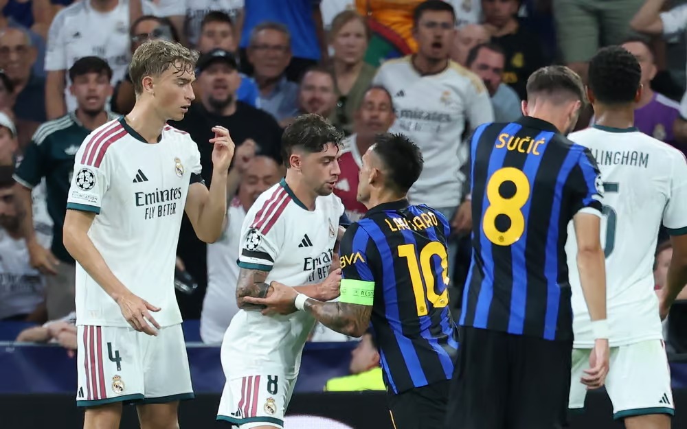 Inter Milan's Lautaro Martínez, in blue-and-black kit, faces off with Real Madrid's Federico Valverde, in white kit, in a tense confrontation on the pitch, with teammates and a visibly reacting crowd around them.