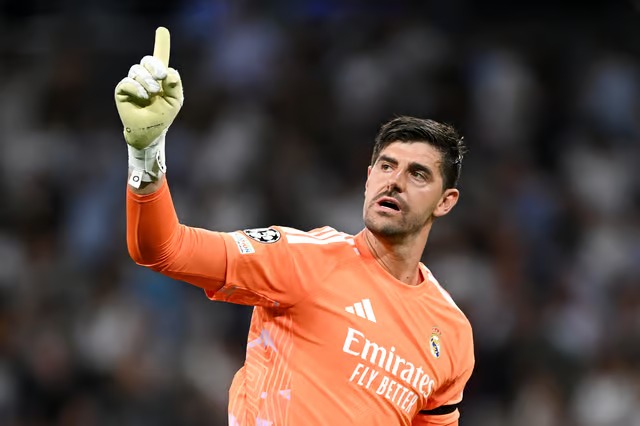 Real Madrid goalkeeper Thibaut Courtois, in an orange kit, raises his gloved hand with index finger pointed up while looking off to the side, with a blurred stadium crowd behind him.