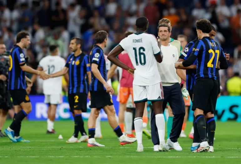 Inter Milan coach Cristian Chivu, in white training gear, exchanges a handshake with Real Madrid defender Ibrahima Konaté (No. 16) on the pitch after the match, with players from both teams greeting each other in the background.