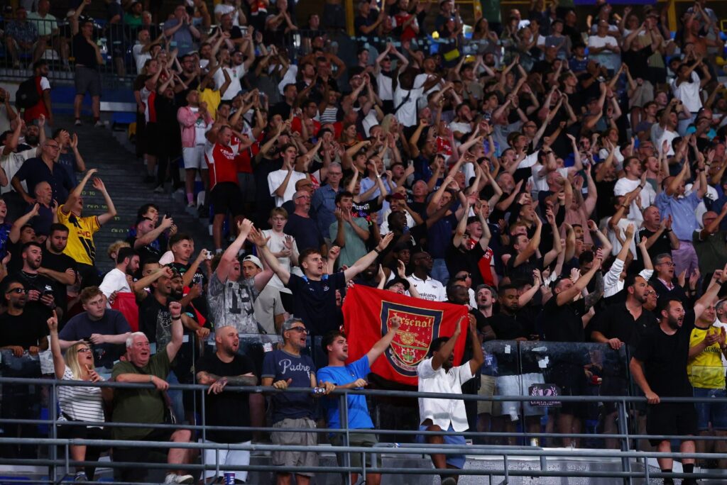 A packed stand of Arsenal fans cheering with arms raised, several wearing club shirts, one holding up a red Arsenal flag with the club crest.