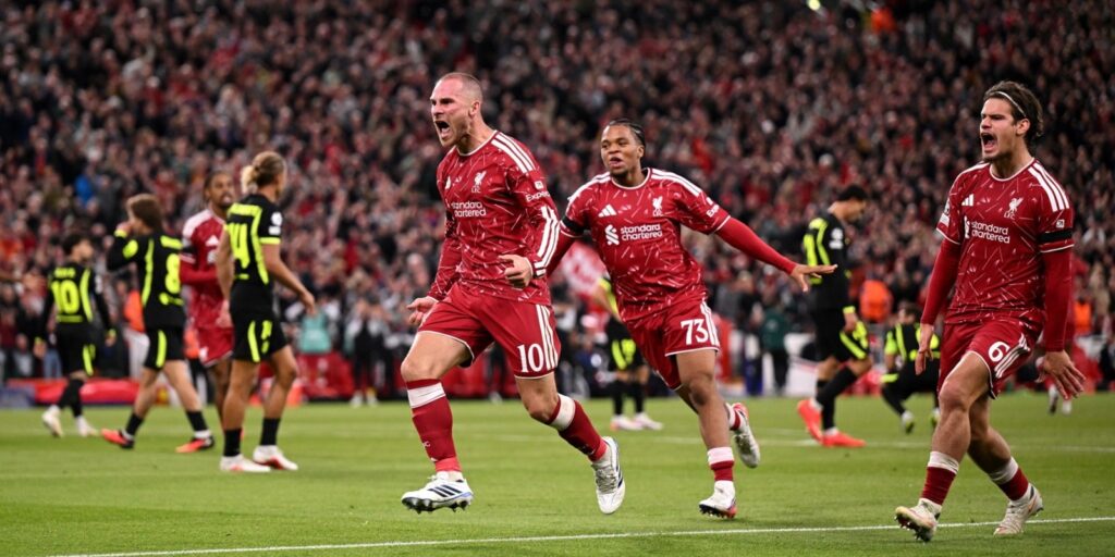 A Liverpool player in a red no. 10 shirt celebrating with arms out and mouth open, chased by teammates including no. 73 and no. 6, opposition players in black and yellow kits in the background, Anfield crowd behind.