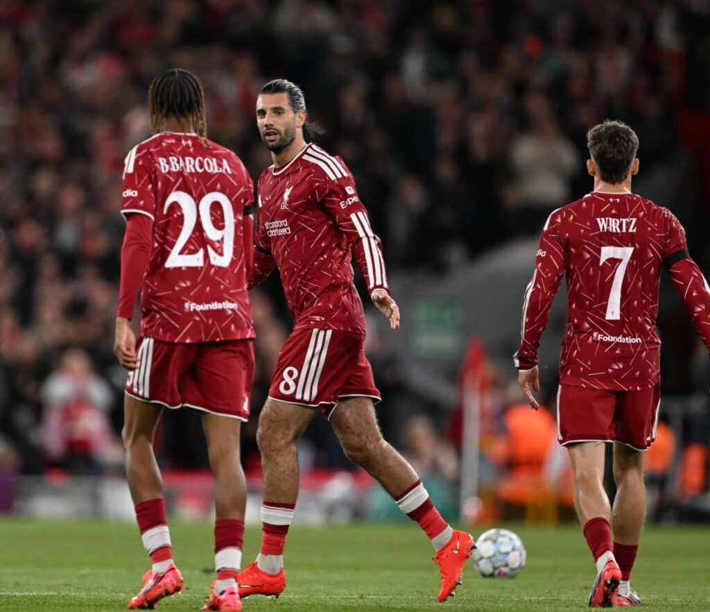 Liverpool players in red kits on the pitch, with visible shirt names "B.Barcola" (No. 29), a No. 8 player facing the camera, and Wirtz (No. 7), a Champions League match ball on the grass nearby.