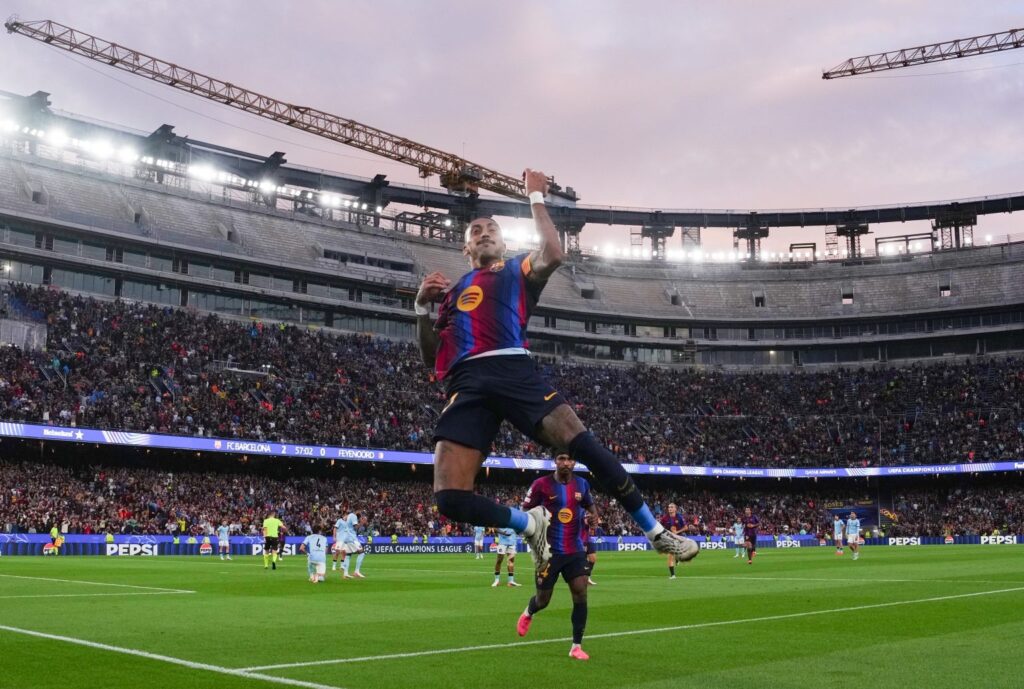 A Barcelona player in a blue-and-maroon kit leaping mid-air in celebration with fist raised, stadium crowd and floodlights in the background, scoreboard reading "FC Barcelona 2 - 0 Feyenoord" at 57:02.