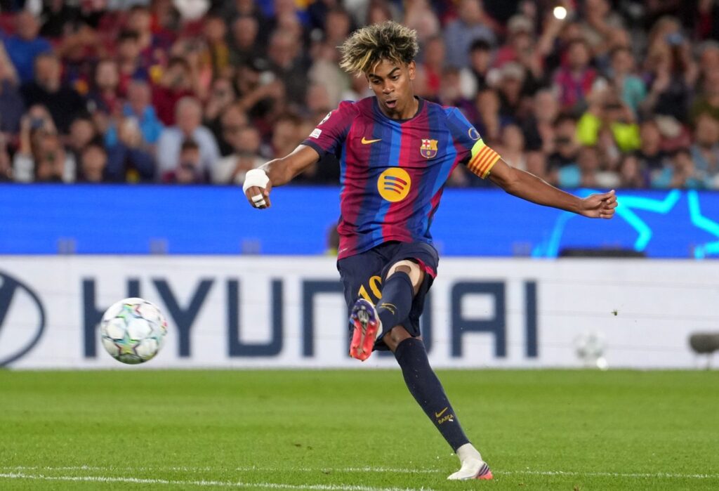 A young Barcelona player in a blue-and-maroon kit following through on a shot, ball in flight, Hyundai advertising board and Champions League ball visible, crowd blurred behind.