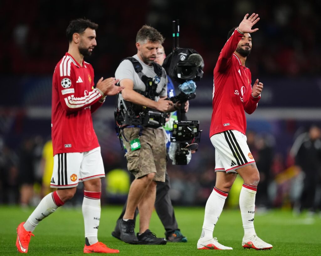Manchester United's Bruno Fernandes and Matheus Cunha applaud fans as they walk off the pitch after the 4-0 win, with a TV cameraman between them.