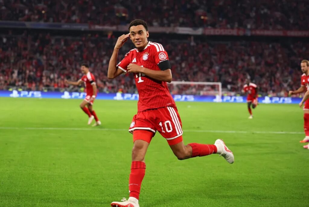 Bayern Munich's Jamal Musiala cups his hand to his ear in celebration after scoring the opening goal in a UEFA Champions League match, as teammates and Harry Kane join in behind him.