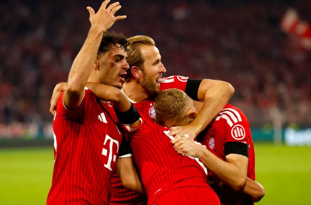 Harry Kane and Bayern Munich teammates embrace in celebration after scoring, huddled together in front of a packed stadium.