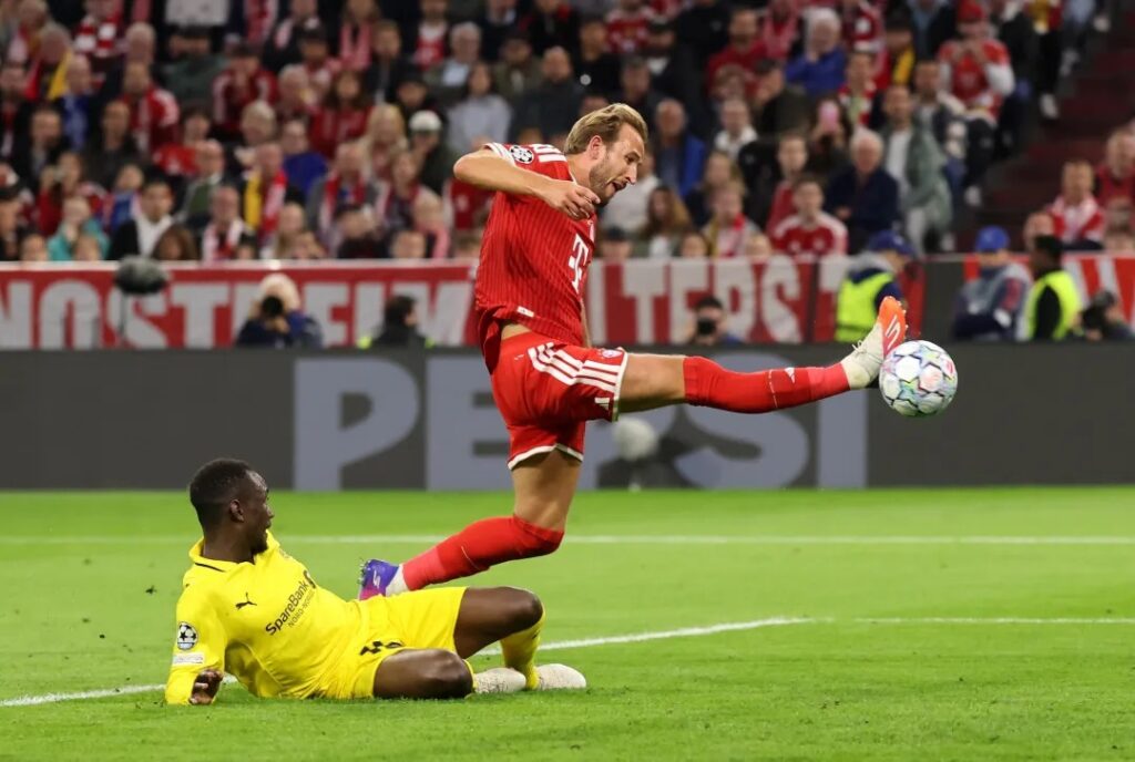 Bayern Munich's Harry Kane leaps to strike the ball past a sliding yellow-shirted defender during a Champions League match.