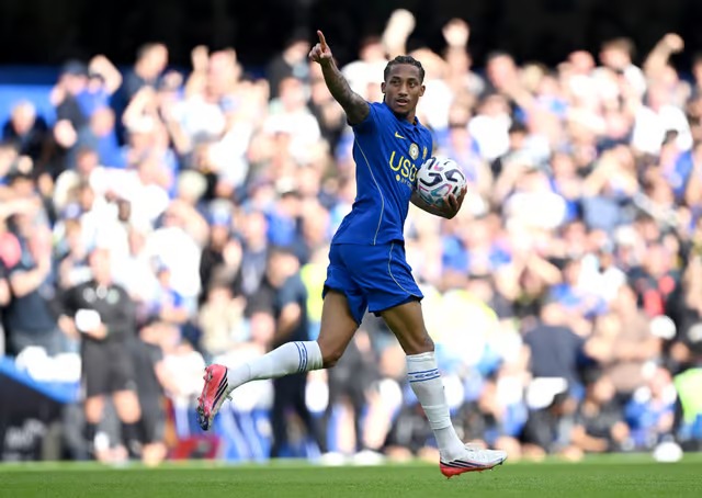 João Pedro of Chelsea celebrating a goal while carrying the match ball, pointing skyward, with jubilant fans in the background at Stamford Bridge.