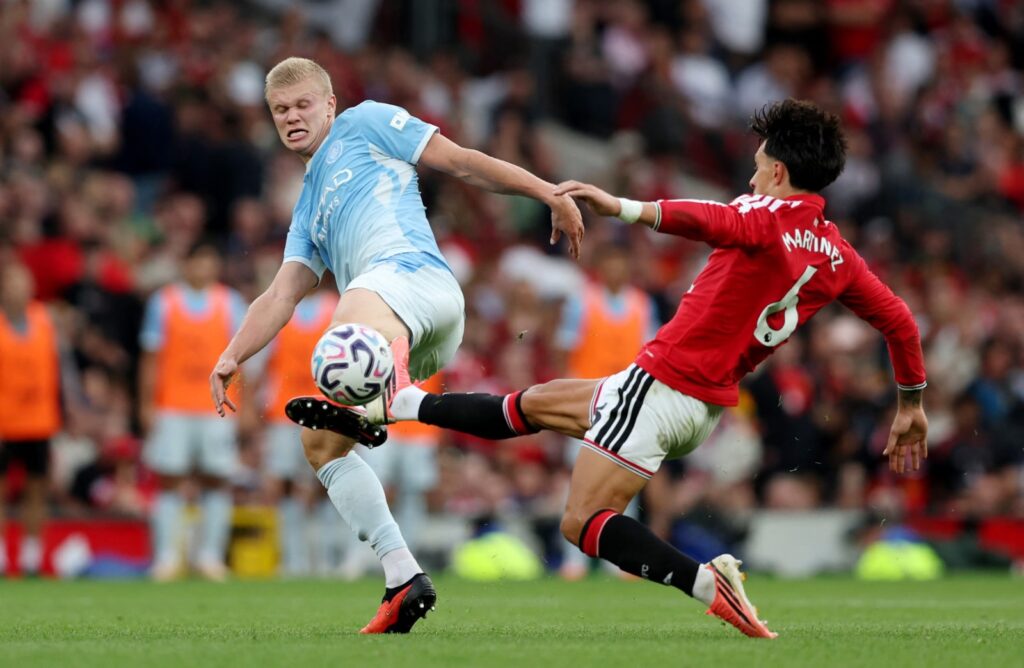 Erling Haaland in Manchester City's sky blue kit contests the ball with Lisandro Martínez of Manchester United in a red shirt marked "Martinez 6", both players stretching for the ball mid-tackle.