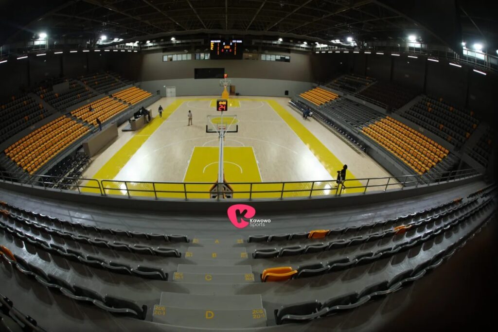 A panoramic view of Hoima City Arena's empty indoor basketball court, with orange and grey tiered seating surrounding the yellow and wood-toned court, taken from a high vantage point.