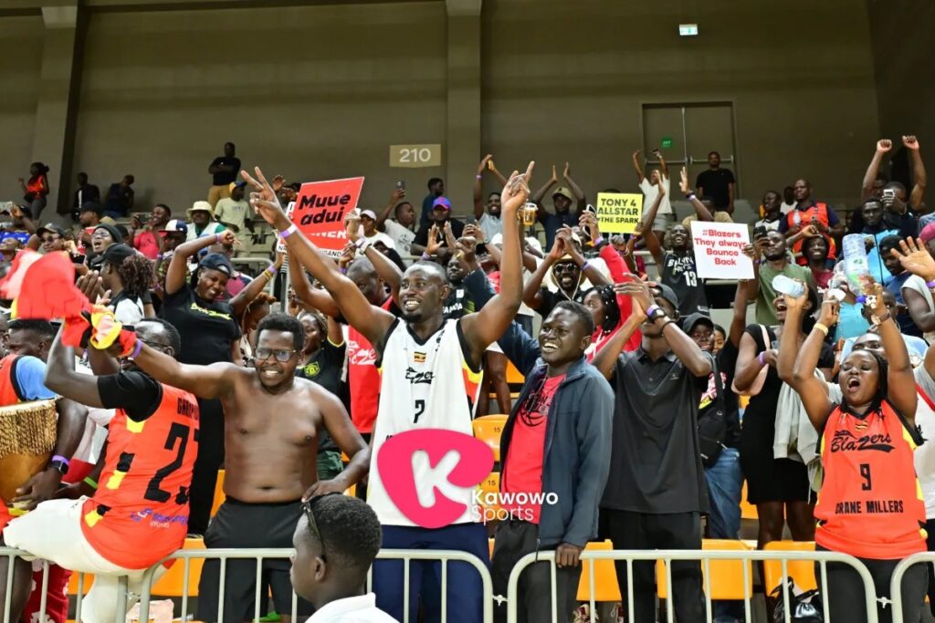 A large crowd of Nam Blazers fans in orange jerseys cheering, waving arms and holding signs including "Blazers They always Bounce Back" in the stands at Hoima City Arena.