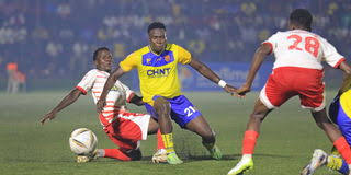 A KCCA FC player in a yellow and blue jersey battles for the ball against Express FC defenders in red and white jerseys during a Uganda Premier League match on a foggy pitch.