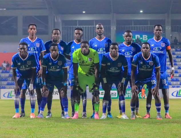 Archive photo of SC Villa football team lined up in two rows on a floodlit pitch, players in blue jerseys and the goalkeeper in green, with stadium stands visible in the background.