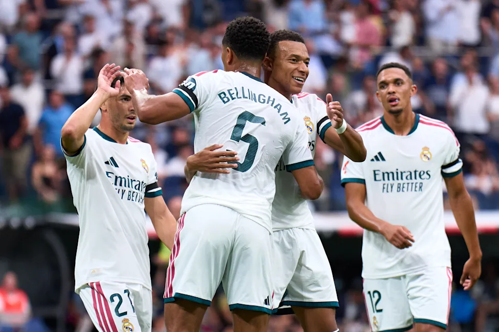 Real Madrid players in white home kits celebrate together on the pitch, embracing near midfielder Jude Bellingham (number 5), with teammates wearing numbers 27 and 12 joining in, and a blurred stadium crowd in the background.