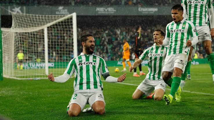 Real Betis players in green-and-white striped kits celebrate a goal on a rain-soaked pitch, with one player sliding on his knees, arms outstretched, while teammates cheer behind him and an opponent looks on near the goalpost.