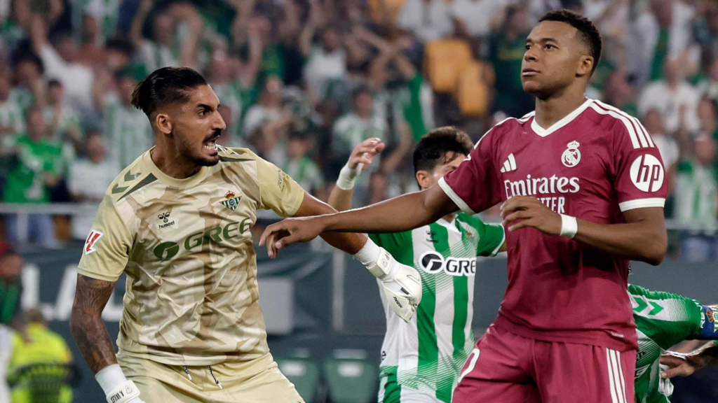 Real Betis goalkeeper reacts angrily and reaches toward Real Madrid's Kylian Mbappé during a tense LaLiga match, with fans visible in the stands behind them.