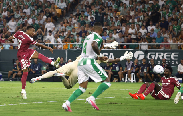 Real Madrid's Kylian Mbappé strikes the ball on goal while the Real Betis goalkeeper dives to block, with defenders and a stadium full of fans in the background.