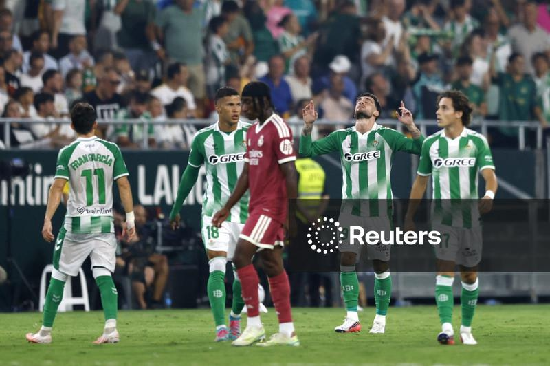 Real Betis players in green and white celebrate a goal, raising their arms, while a Real Madrid player in maroon looks down dejectedly nearby.