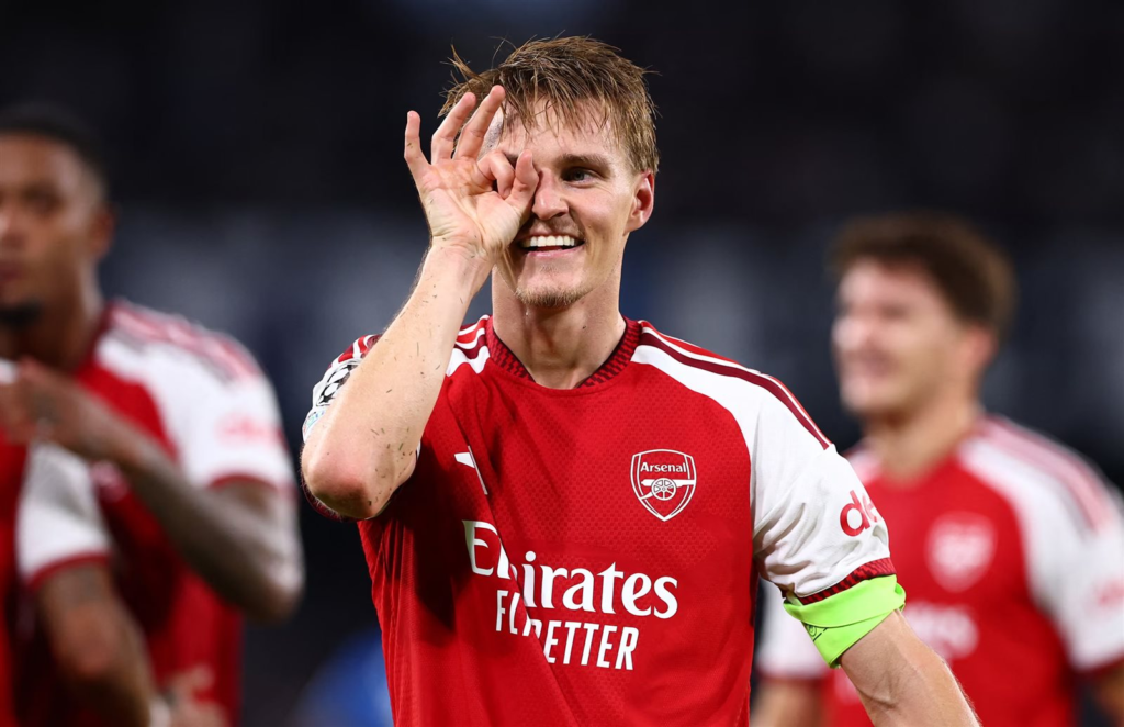 Arsenal captain Martin Ødegaard, wearing the captain's armband, celebrates with his signature eye-rubbing gesture, teammates blurred in the background.