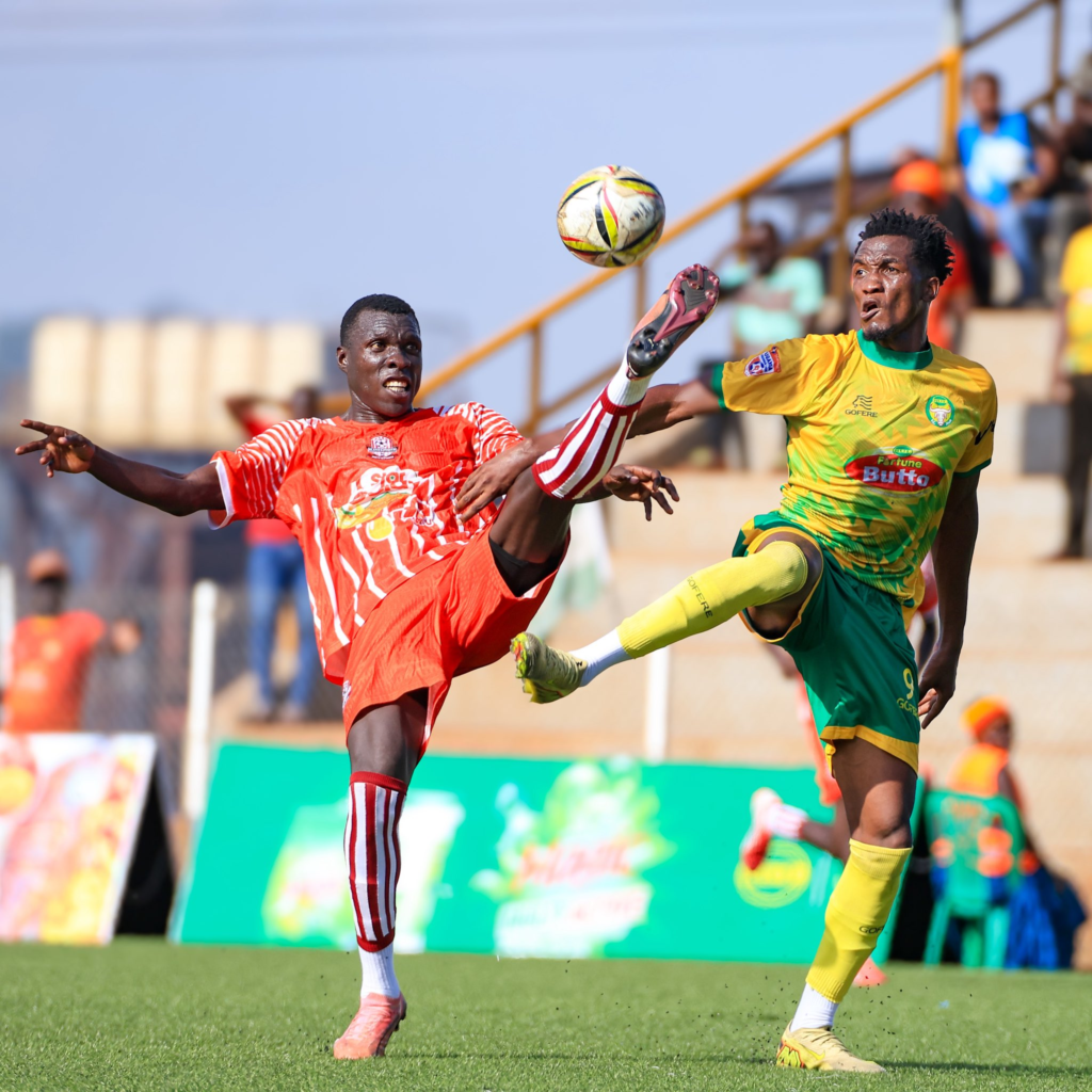 A Blacks Power player in red and white and a BUL FC player in yellow and green stretch to control a ball in the air during a match.