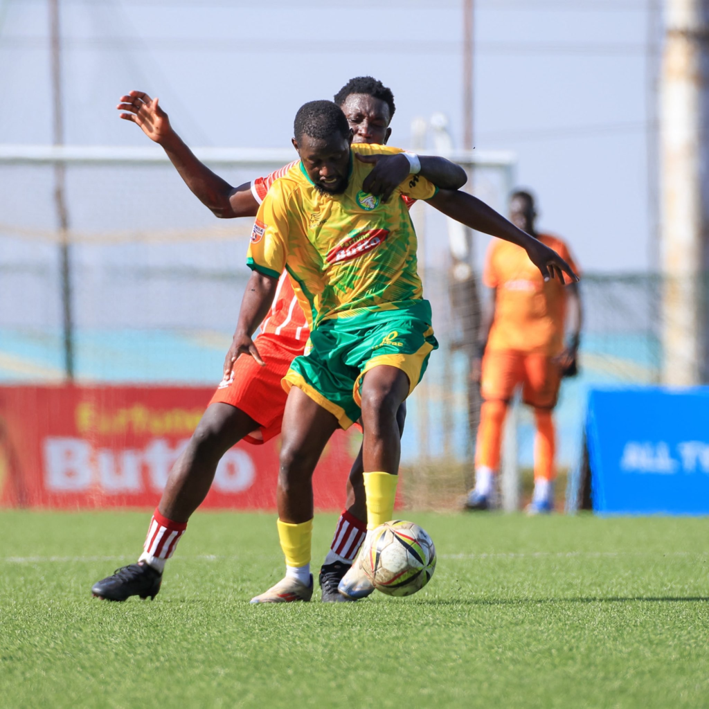 A BUL FC player in yellow and green shields the ball from a Blacks Power player in orange and red during a match.